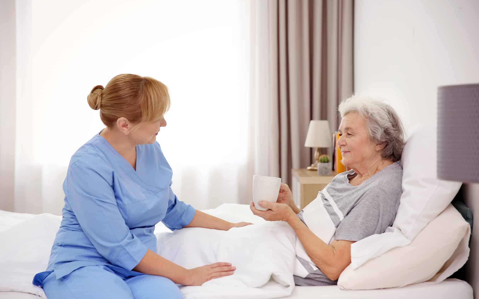 Female caregiver checking in with an elderly woman as she rests in her bed