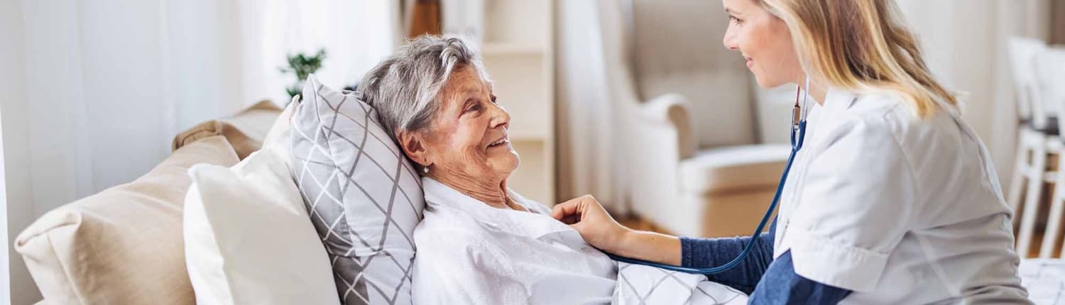 A health visitor examining a sick senior woman lying in bed at home with stethoscope_